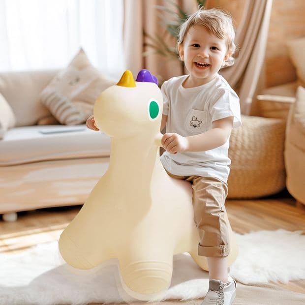 Toddler in a bright living room standing and holding onto a cream inflatable dinosaur hopper, mid-laugh