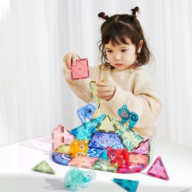 Young girl at a white table placing a pink translucent magnetic tile onto a forest-animal tile build