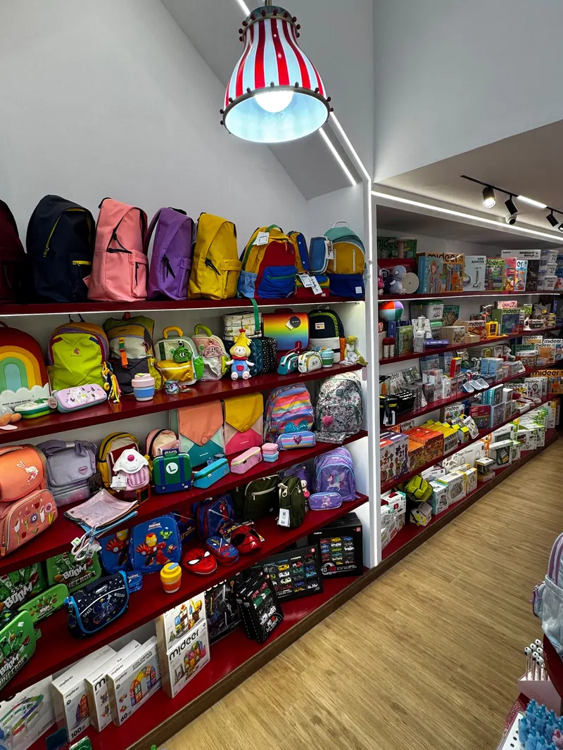 A wall of children's backpacks on red shelves under a red and white striped pendant lamp, inside a white house-shaped frame.
